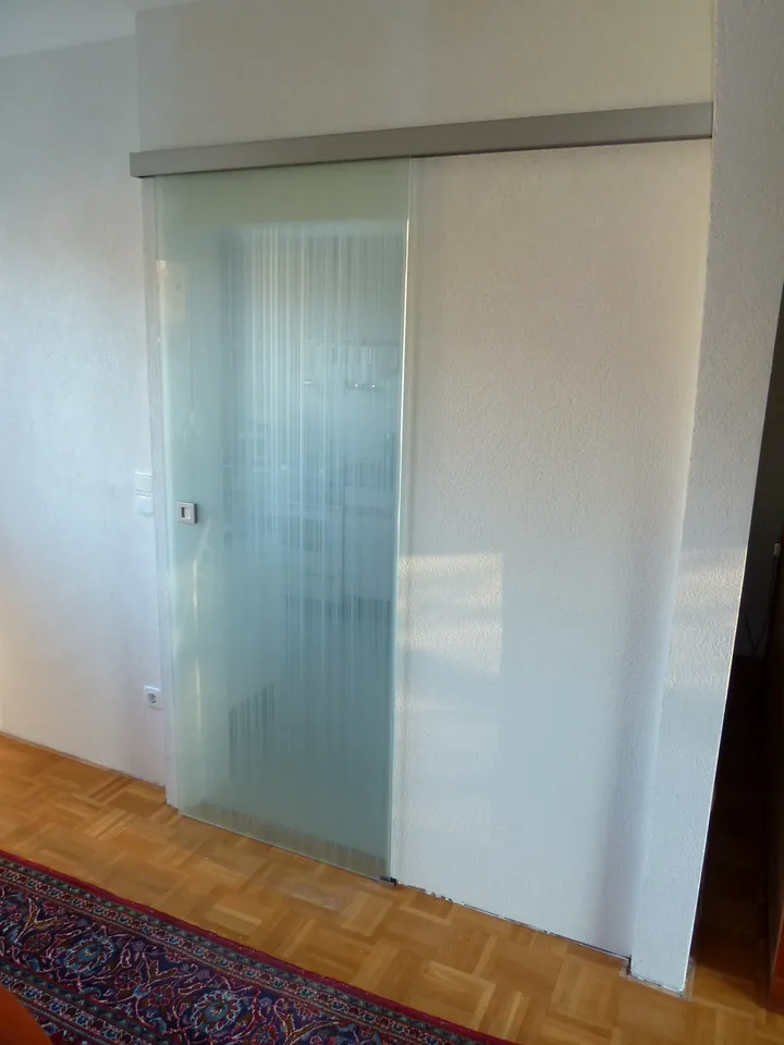 Glasschiebetür mit dezentem Streifenmuster und oberer Laufschiene vor einer weißen Wand